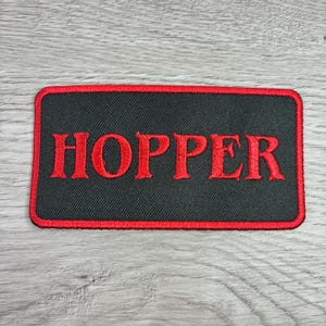 May include: A rectangular patch with the name "HOPPER" embroidered in red on a black background. The patch has a red border and is likely designed to be sewn or ironed onto clothing or accessories.