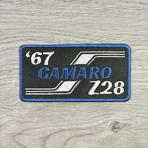 May include: Black rectangular patch with a blue border. The patch features the text "'67 CAMARO Z28" in white and blue. The design includes white stripes and is likely an emblem for a classic car.