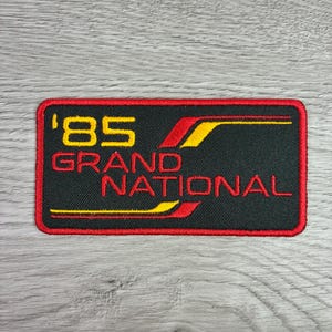 May include: Embroidered patch with a black background and a red border. The patch features the text "'85 GRAND NATIONAL" in yellow and red. A yellow and red design element is on the right side of the patch.