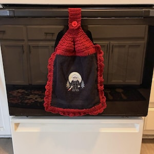May include: A dark gray hanging kitchen towel featuring a red crocheted top with a matching red button closure. The towel has a red crocheted border and an embroidered graphic of a hockey-masked character holding a knife in front of a moon.