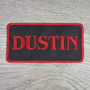 May include: A rectangular embroidered patch with a black background and a red border. The word DUSTIN is stitched in large, bold, red capital letters across the center of the patch. The design features a clean, uniform texture throughout the surface.