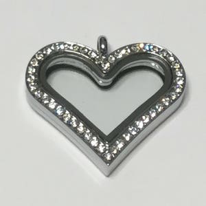 Könnte beinhalten: Ein silberner herzförmiger Anhänger mit einem klaren Glaseinsatz und einer doppelten Reihe von klaren Strasssteinen am Rand.