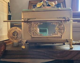 May include: A gold-coloured decorative box with ornate detailing, featuring a mirrored front panel and decorative legs. A figurine of a man in a hat stands on top. The box has gold handles and a circular emblem on the side.
