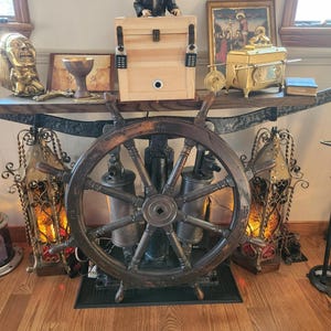 Puede incluir: Una timonera de madera oscura es el punto focal de la imagen. Detr&aacute;s, una exhibici&oacute;n decorativa incluye una caja de madera con cerraduras de combinaci&oacute;n, una figura y varios objetos antiguos. La escena se desarrolla sobre un suelo de madera.