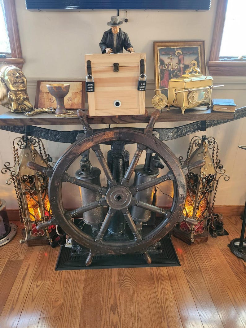 Puede incluir: Una timonera de madera oscura es el punto focal de la imagen. Detr&aacute;s, una exhibici&oacute;n decorativa incluye una caja de madera con cerraduras de combinaci&oacute;n, una figura y varios objetos antiguos. La escena se desarrolla sobre un suelo de madera.