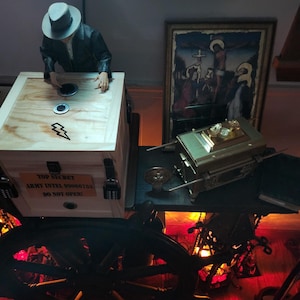 Puede incluir: Una caja de madera con el texto "TOP SECRET ARMY INTEL 99066753 DO NOT OPEN!" est&aacute; junto a una figura con sombrero. Una caja dorada y una pintura enmarcada est&aacute;n sobre una mesa. Una rueda grande y oscura est&aacute; en primer plano.