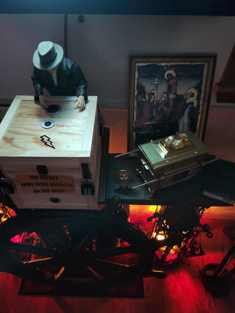 Puede incluir: Una caja de madera con el texto "TOP SECRET ARMY INTEL 99066753 DO NOT OPEN!" est&aacute; junto a una figura con sombrero. Una caja dorada y una pintura enmarcada est&aacute;n sobre una mesa. Una rueda grande y oscura est&aacute; en primer plano.