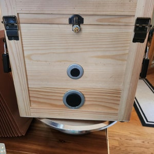 Puede incluir: Una caja de madera de color claro con tapa con bisagras y herrajes met&aacute;licos. La caja presenta una cerradura, dos orificios de ventilaci&oacute;n circulares y pestillos met&aacute;licos. La veta de la madera es visible.