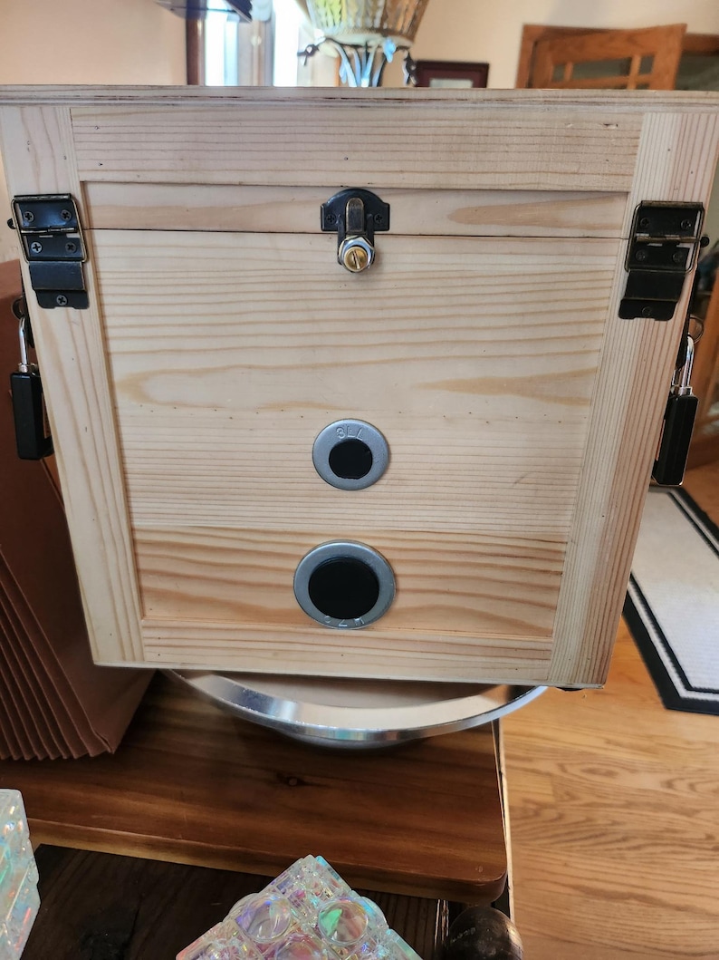 Puede incluir: Una caja de madera de color claro con tapa con bisagras y herrajes met&aacute;licos. La caja presenta una cerradura, dos orificios de ventilaci&oacute;n circulares y pestillos met&aacute;licos. La veta de la madera es visible.