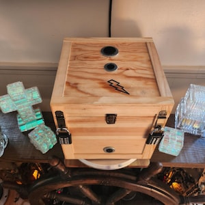Puede incluir: Una caja de madera natural con un rayo negro. La caja tiene dos aberturas circulares en la parte superior y una cerradura de combinaci&oacute;n. Piezas de vidrio decorativas y un tim&oacute;n est&aacute;n en el fondo.