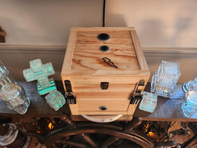 Puede incluir: Una caja de madera natural con un rayo negro. La caja tiene dos aberturas circulares en la parte superior y una cerradura de combinaci&oacute;n. Piezas de vidrio decorativas y un tim&oacute;n est&aacute;n en el fondo.