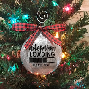 May include: Silver glitter Christmas ornament with a black and red buffalo plaid ribbon. The ornament has the text "adoption loading" with a progress bar and the words "please wait".