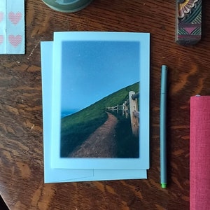 May include: A greeting card with a landscape photo of a path on a green hillside under a starry sky. A green mug with coffee, a wooden cross, a pen, and a red book are on a wooden table.