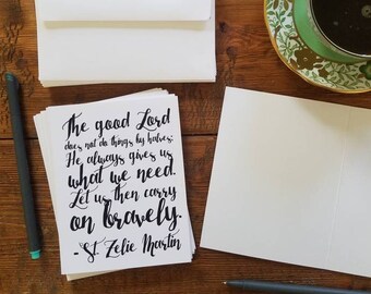 May include: A collection of stationery and a cup of coffee arranged on a wooden table. The image shows white envelopes, cards with a handwritten quote, a blank card, two pens, and a teacup with a floral pattern.