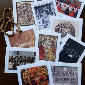 May include: A selection of square greeting cards displaying religious art, illustrating biblical scenes. The cards showcase diverse styles, including colour paintings and monochrome etchings. A wooden rosary with a cross is also visible.