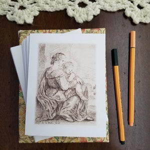 May include: A stack of art prints featuring a sepia-toned illustration of a woman and child, resting on a decorative box. Two orange pens with black caps are placed to the right. A cream-colored crocheted lace doily is at the top.