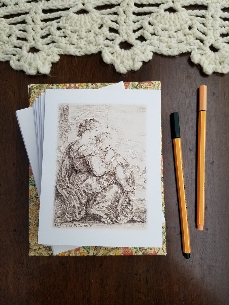 May include: A stack of art prints featuring a sepia-toned illustration of a woman and child, resting on a decorative box. Two orange pens with black caps are placed to the right. A cream-colored crocheted lace doily is at the top.