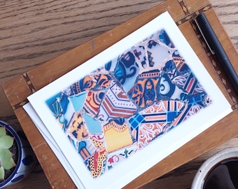 May include: A stack of greeting cards featuring a mosaic tile design in shades of blue, orange, and white. A black pen rests on the wooden frame holding the cards. A small succulent in a blue and white pot and a cup of coffee are also in the image.