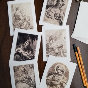 May include: A collection of art print greeting cards featuring sepia-toned illustrations of religious figures. The cards depict scenes of mothers and children, with detailed linework and a vintage aesthetic. A stack of blank cards and pens are also visible.