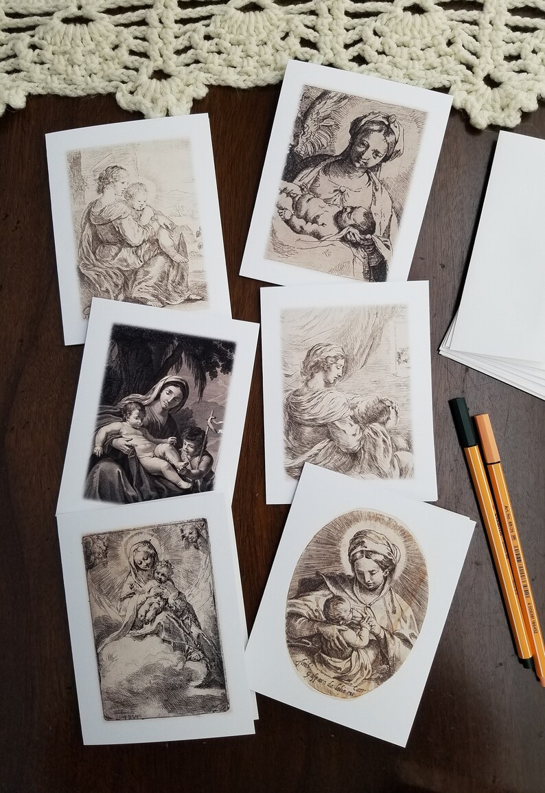 May include: A collection of art print greeting cards featuring sepia-toned illustrations of religious figures. The cards depict scenes of mothers and children, with detailed linework and a vintage aesthetic. A stack of blank cards and pens are also visible.