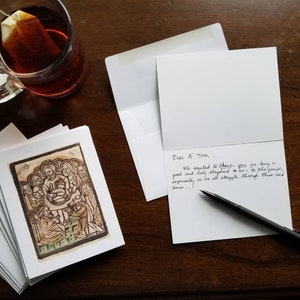 May include: A collection of white greeting cards with envelopes, one card open with handwritten text. The cards display a sepia-toned religious illustration. A glass mug of tea and a black pen are on a dark wooden surface.