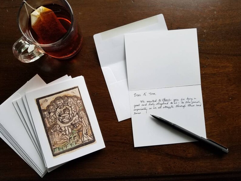 May include: A collection of white greeting cards with envelopes, one card open with handwritten text. The cards display a sepia-toned religious illustration. A glass mug of tea and a black pen are on a dark wooden surface.