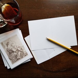 May include: A stack of blank greeting cards with a pencil and envelope on a dark wooden surface. A glass mug of tea and a stack of cards with a sepia-toned illustration are also present.