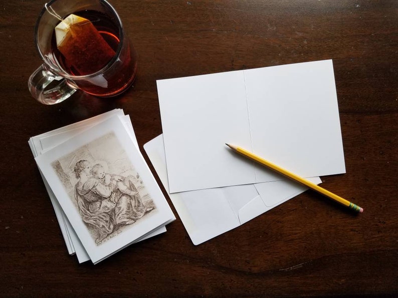 May include: A stack of blank greeting cards with a pencil and envelope on a dark wooden surface. A glass mug of tea and a stack of cards with a sepia-toned illustration are also present.