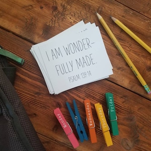 May include: A stack of white cards with the text "I AM WONDERFULLY MADE. PSALM 139:14" sits on a wooden surface, next to two yellow pencils and colorful clothespins labeled with weekdays. A dark green bag is in the background.
