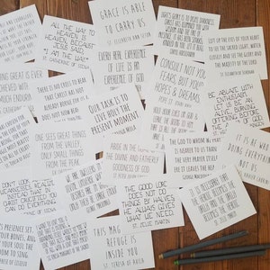 May include: A selection of white cards with black text, displaying inspirational quotes and religious sayings. The cards are arranged on a wooden surface, with several pens nearby. The quotes are from various religious figures.