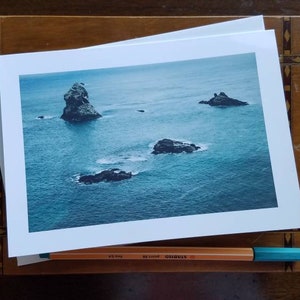 May include: A stack of landscape art prints featuring a seascape with rock formations in the ocean. The prints are on a wooden box with decorative inlay. A pen with a teal cap rests on the box.