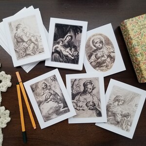 May include: A collection of religious art greeting cards featuring black and white illustrations of Madonna and Child. The cards are displayed on a dark wooden surface, alongside a decorative box and two pens. The cards are blank inside.