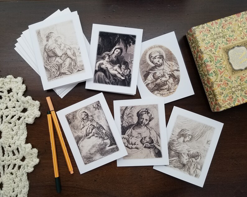 May include: A collection of religious art greeting cards featuring black and white illustrations of Madonna and Child. The cards are displayed on a dark wooden surface, alongside a decorative box and two pens. The cards are blank inside.