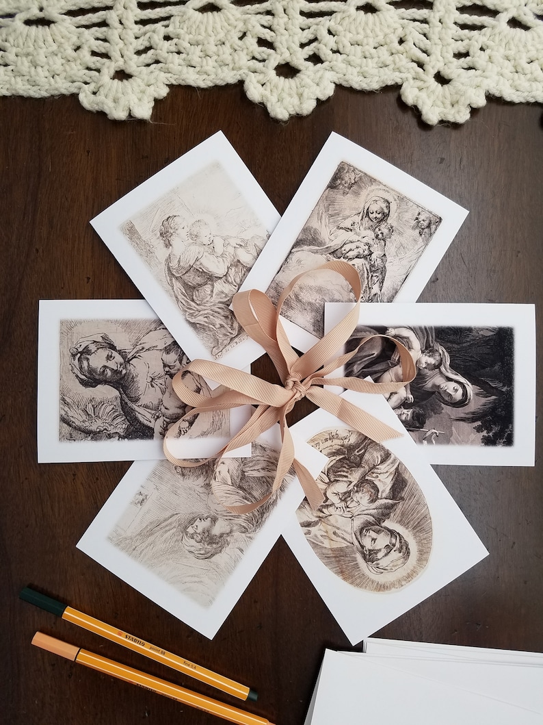 May include: A collection of art prints featuring religious figures, tied with a tan ribbon. The prints are in black and white, with detailed illustrations. Two orange and green pens are visible, along with a white lace doily.