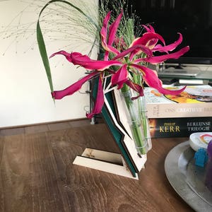Op de afbeelding: Een houten bloemenstandaard met een glazen vaas met roze en gele bloemen. De standaard is gemaakt van drie stukken hout, elk met een andere kleur en patroon. De standaard staat op een houten tafel met een boek en een bord erop.