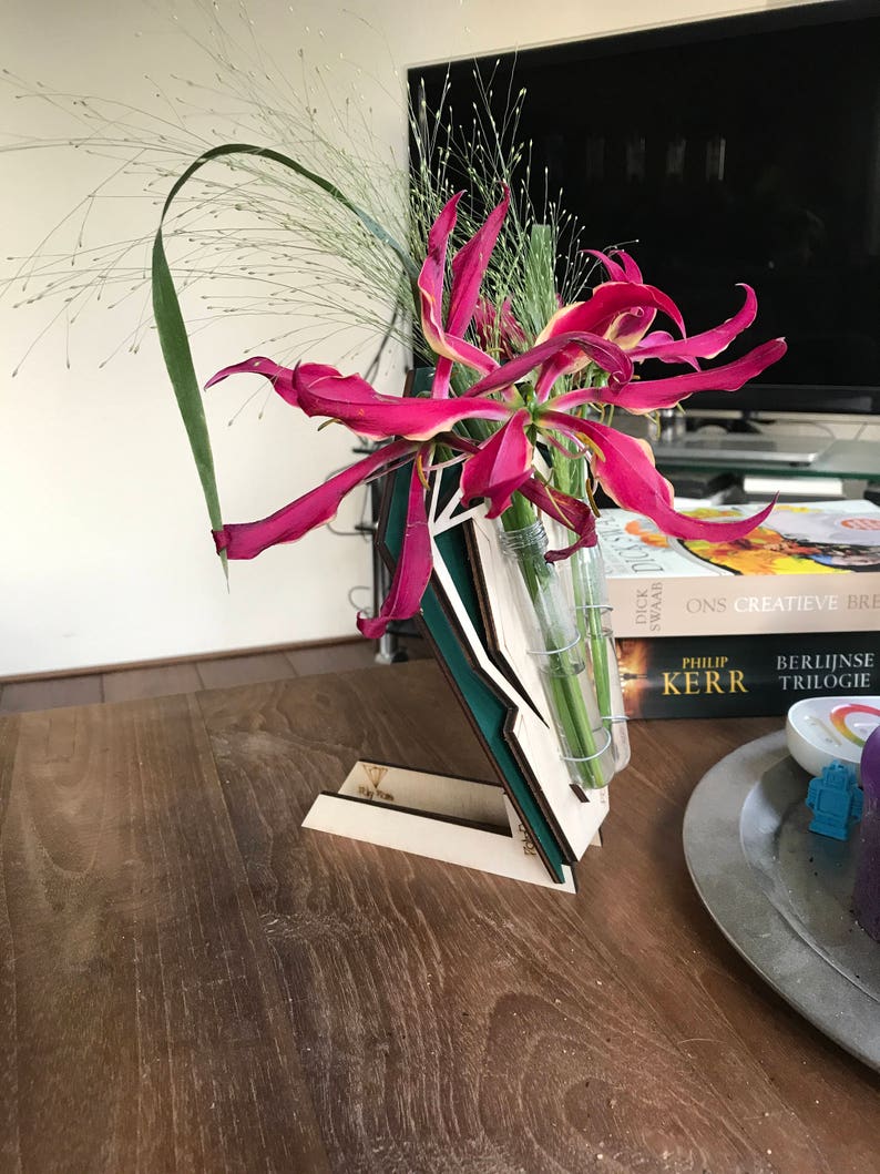 Op de afbeelding: Een houten bloemenstandaard met een glazen vaas met roze en gele bloemen. De standaard is gemaakt van drie stukken hout, elk met een andere kleur en patroon. De standaard staat op een houten tafel met een boek en een bord erop.