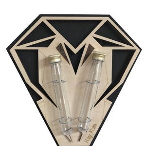 Op de afbeelding: Een houten diamantvormige wandbevestiging met twee glazen reageerbuizen en gouden deksels. De bevestiging is zwart met een houtnerfpatroon en de tekst "Poly Rare" is onderaan gedrukt.