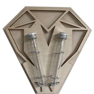 Op de afbeelding: Een houten diamantvormige wanddecoratie met twee heldere glazen reageerbuizen met zilveren deksels. De reageerbuizen worden op hun plaats gehouden door draadringen.