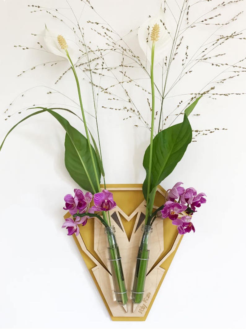 Op de afbeelding: Een houten wandvaas met twee glazen buizen om bloemen in te steken. De vaas is geel geverfd en heeft een geometrisch ontwerp. De vaas bevat roze orchidee&euml;n en witte bloemen.