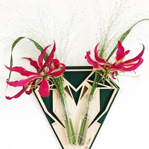 Op de afbeelding: Een groene geometrische houten wandvaas met twee glazen buizen om bloemen in te steken. De vaas heeft de tekst "Toby Ross" op de bodem. De vaas bevat roze bloemen en groene stelen.
