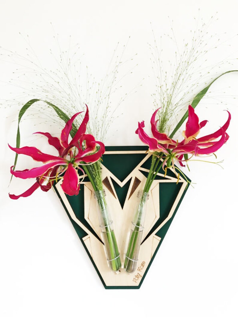 Op de afbeelding: Een groene geometrische houten wandvaas met twee glazen buizen om bloemen in te steken. De vaas heeft de tekst "Toby Ross" op de bodem. De vaas bevat roze bloemen en groene stelen.