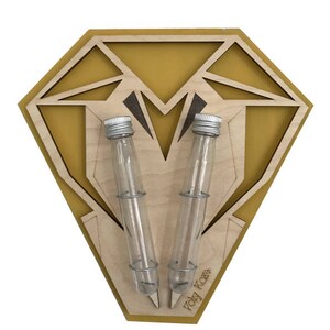 Op de afbeelding: Een houten wandrek voor kruiden in de vorm van een diamant met een gele achtergrond. Twee glazen reageerbuizen met zilveren deksels zijn in het rek gestoken. Het rek is gelabeld met "Toby Fox".