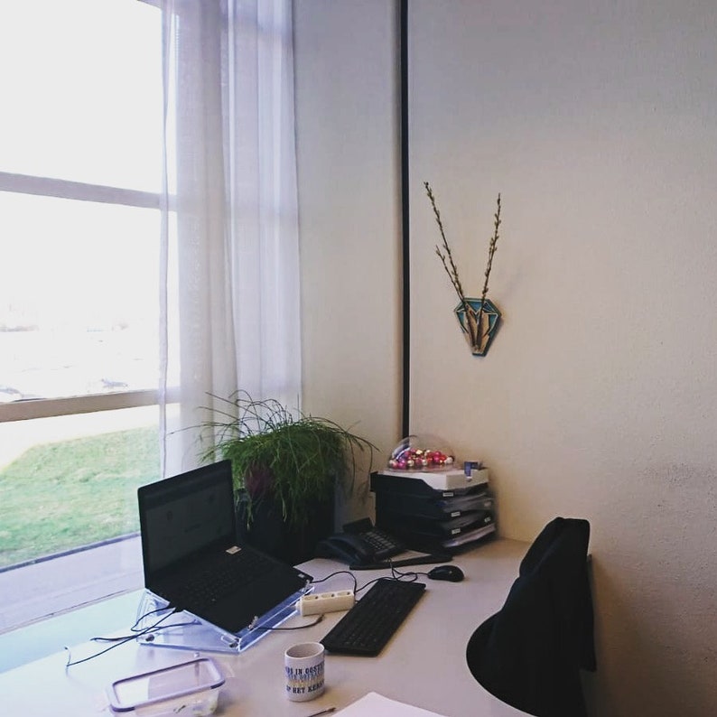 Op de afbeelding: Een wit bureau met een laptop, een toetsenbord, een mok en een doorzichtige plastic container. Er staat een plant in een pot op het bureau. Het bureau staat naast een raam met witte gordijnen. Er staat een zwarte stoel op de achtergrond.