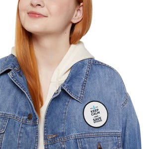 Op de afbeelding: Een lichtblauwe spijkerjas met een witte hoodie eronder. Een ronde patch op de jas leest "KEEP CALM AND SAVE SPOONS" met een turquoise lintafbeelding. De jas heeft een knoopsluiting en zakken.