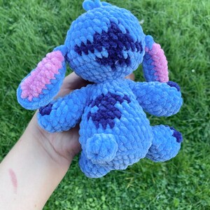 May include: A handmade blue crochet plush toy with dark blue and pink accents. The plushie has large ears, a round body, and small arms and legs. The toy is held in a hand against a green grass background.