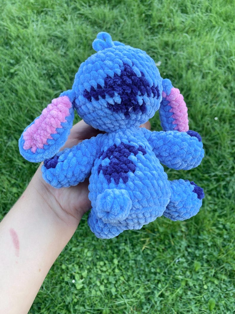 May include: A handmade blue crochet plush toy with dark blue and pink accents. The plushie has large ears, a round body, and small arms and legs. The toy is held in a hand against a green grass background.