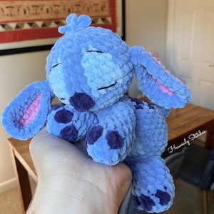May include: A blue plush toy character with large ears, purple spots, and closed eyes. The handmade stuffed animal is a soft, cuddly toy. The ears have pink inner details. The toy is held in a person's hand.