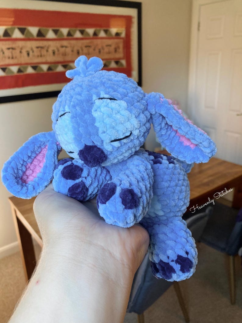 May include: A plush blue character with large ears, purple spots, and closed eyes. The handmade stuffed animal is a soft, cuddly toy. The ears have pink inner details. The toy is held in a person's hand.