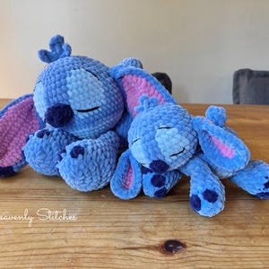 May include: Two blue crochet plush toys resembling a cartoon character, with large ears and closed eyes, are resting on a wooden surface. The toys are in shades of blue with pink accents on the ears. The text "Heavenly Stitches" is visible.
