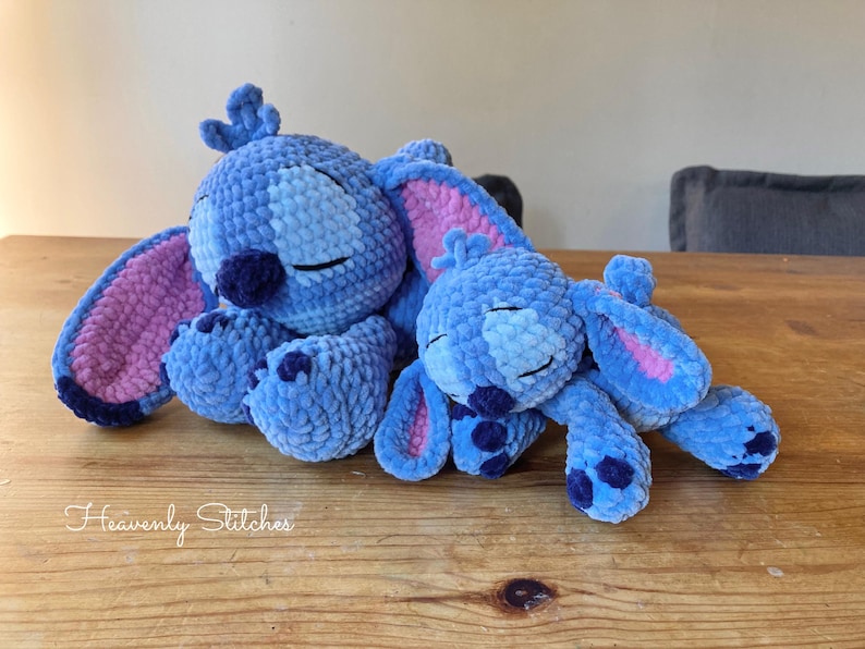 May include: Two blue crochet plush toys resembling a cartoon character, with large ears and closed eyes, are resting on a wooden surface. The toys are in shades of blue with pink accents on the ears. The text "Heavenly Stitches" is visible.
